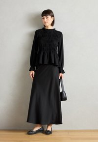 Černá peplum blůza s řasenou strukturou, dlouhými rukávy a volánkovým výstřihem, spárovaná s černou maxi sukní a plochými botami s mašlí.