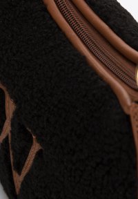 Sac en polaire noir avec fermeture éclair et accents en cuir brun. Surface texturée avec un motif subtil, présentant des découpes triangulaires d'un côté.