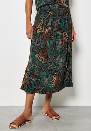 Femme portant une jupe midi à motifs de feuilles vertes, brunes et noires, avec des sandales plates ouvertes marron, debout sur un sol gris.