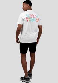 Camiseta blanca de algodón con el texto colorido "AMIGOS INCORRECTOS" goteando en la parte posterior, combinada con pantalones cortos negros y zapatillas deportivas grises.