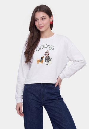 Donna con lunghi capelli castani che indossa una felpa bianca corta con illustrazioni di cani e testo "Cute Dogs,", abbinata a pantaloni blu scuro.