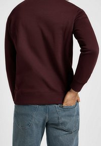 Persona che indossa una camicia a maniche lunghe color bordeaux e jeans blu, con una mano nella tasca posteriore, vista da dietro su uno sfondo bianco.