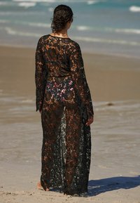 Copertura in pizzo nero con maniche lunghe, motivo floreale, design trasparente e apertura frontale. Indossata sopra un costume da bagno stampato con fiori.