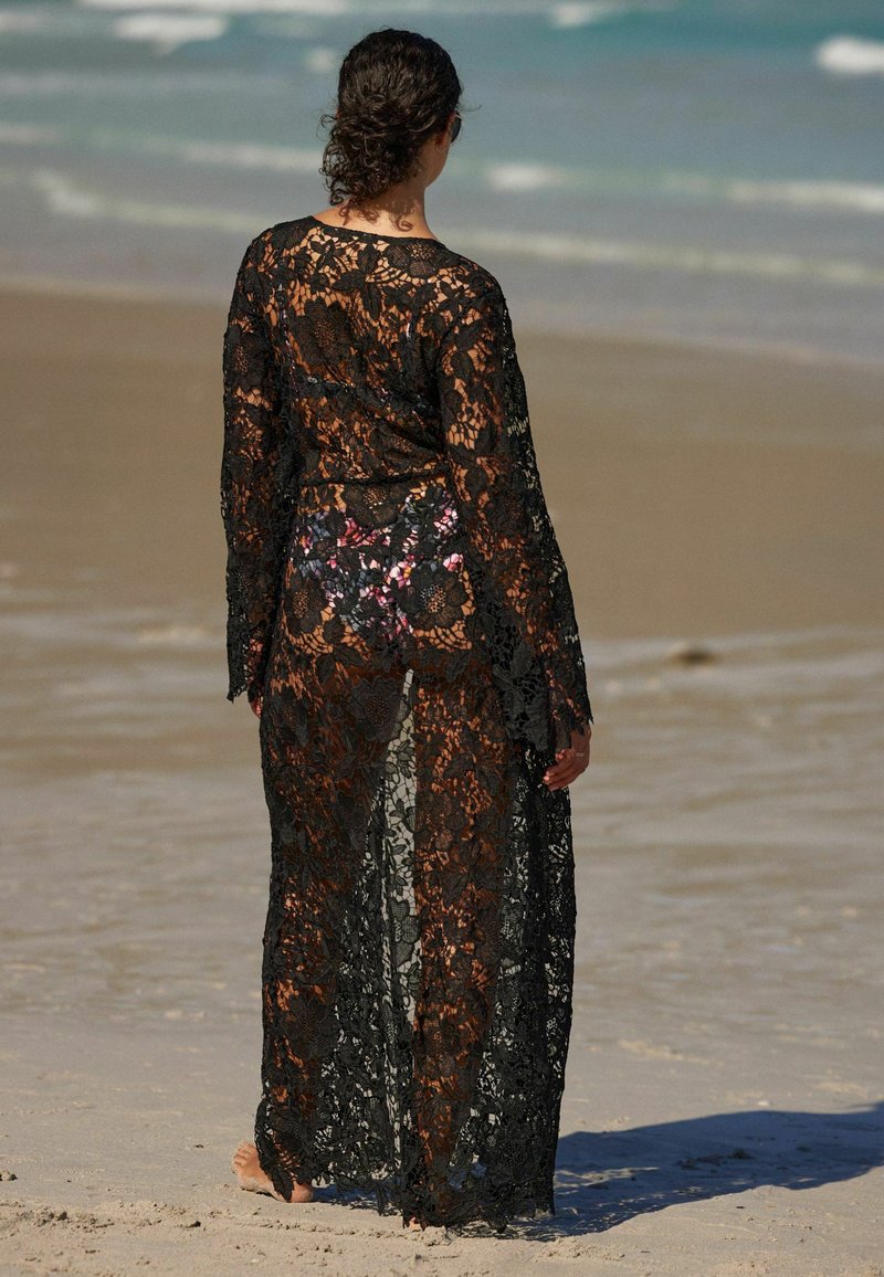 Copertura in pizzo nero con maniche lunghe, motivo floreale, design trasparente e apertura frontale. Indossata sopra un costume da bagno stampato con fiori.