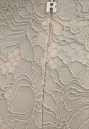 Tessuto beige con sovrapposizione di pizzo bianco intricato che presenta motivi floreali e una fine texture a rete, fissato con una spilla sormontata dalla lettera "R" in argento.