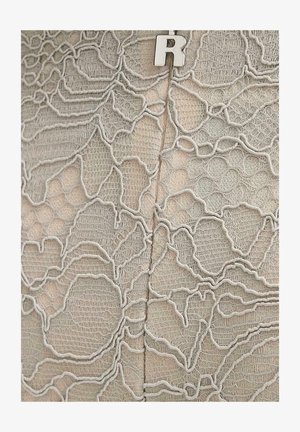 Tissu beige avec une superposition délicate de dentelle blanche présentant des motifs floraux et une texture fine en maille, maintenue par une épingle surmontée d'une lettre "R" en argent.