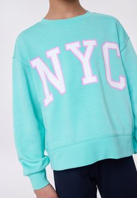 Pullover in turchese chiaro con lettering bianco e rosa "NYC". Presenta polsini e orlo a coste, realizzato in morbido cotone.