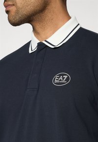 Μπλε ναυτικό πόλο με γιακά ριγέ σε λευ κό και μπλε. Διαθέτει κεντητό λογότυπο EA7 στο στήθος. Κατασκευασμένο από υφασμάτινη υφή.
