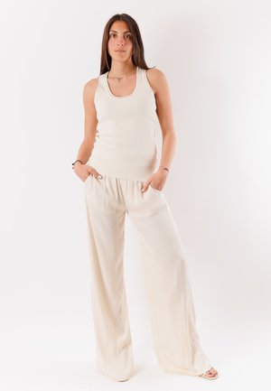 Donna con lunghi capelli scuri che indossa un top bianco senza maniche e pantaloni crema a gamba larga, in piedi con le mani in tasca su sfondo bianco.