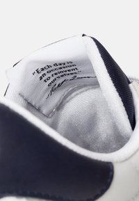 Wnętrze białego buta sportowego z granatowymi akcentami. Miękka tekstylna wyściółka; metka zawiera wydrukowany czarny cytat. Wyściełanie wokół kołnierza.