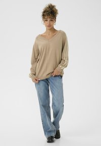Beige V-Ausschnitt Strickpullover mit langen Ärmeln, der ein helleres Streifenmuster auf den Ärmeln aufweist; kombiniert mit blauen weiten Jeans und schwarzen Schuhen.