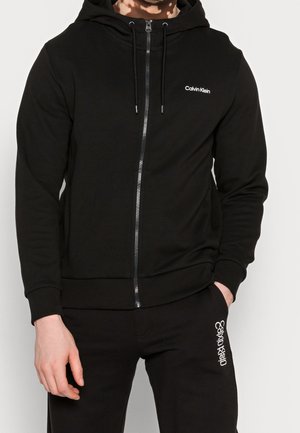 Sudadera negra con cremallera hecha de un tejido suave, con cremallera frontal, capucha y el logo de Calvin Klein en el pecho. Cuerdas ajustables.