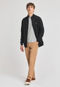 Svart zip-up jacka över en grå t-shirt, kombinerad med tan-färgade byxor och bruna sneakers. Mjukt material, avslappnad design och figurnära passform.