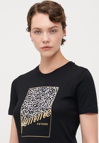 Svart t-skjorte med leopardmønster og gullakenter med teksten "femme FUTURE." Korte ermer og klassisk rund hals design.