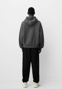 Grå huvtröja med en avslappnad passform, ribbade muddar och en kenguruficka, tillsammans med svarta löst sittande byxor och ljusa skor.