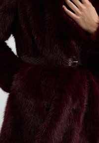 Käsi puusalla, syvän burgundin värisessä keinoturkissa, jossa on teksturoitu vastaava vyö ja kultainen solki.