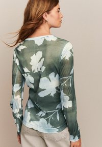 Kvinna sedd bakifrån som bär en grön långärmad topp med stora vita blommöster och beige byxor, hår som blåser lätt.