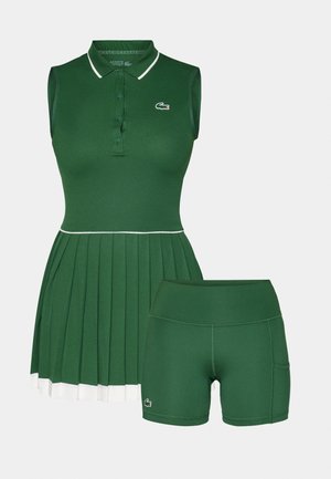 Zöld ujjatlan pólóruha plissé szoknyával és fehér részletekkel, zöld összeillő sorttal párosítva. A sortokon a Lacoste logó látható.