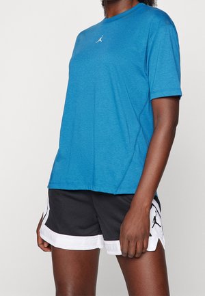 Persoon draagt een blauw T-shirt met korte mouwen en zwarte sportshorts met witte bies, voorzien van een klein wit logo op beide kledingstukken.