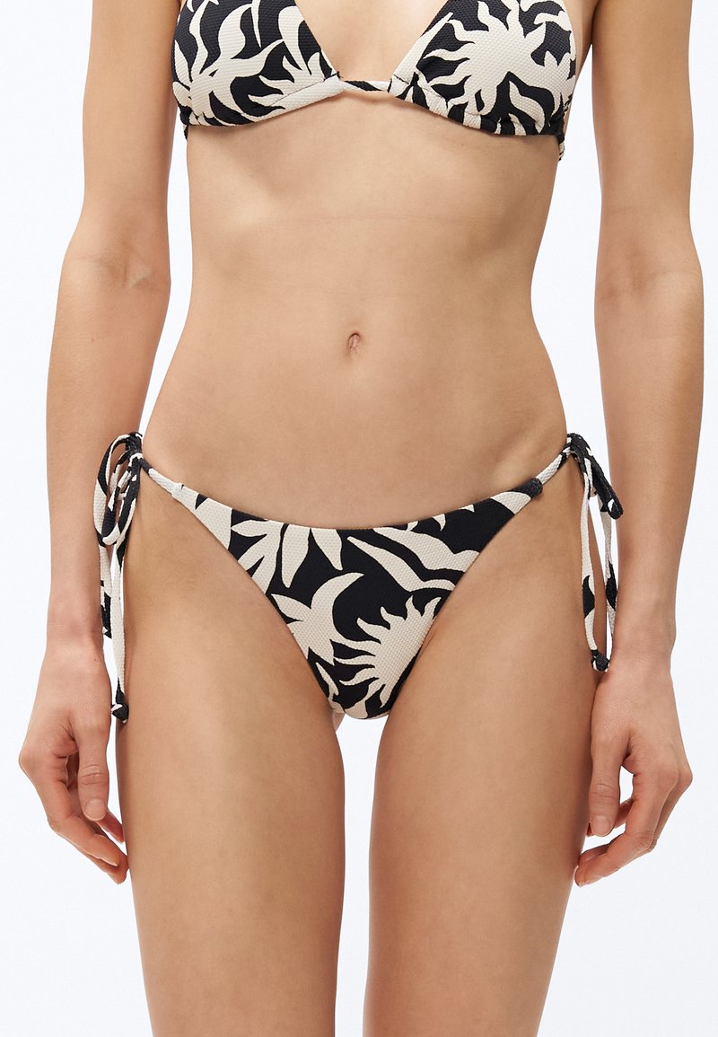 Nő fekete-fehér, oldalt megkötős bikini alsóban, botanikai mintákkal, egyszínű háttér előtt, a törzsét és csípőjét mutatva.