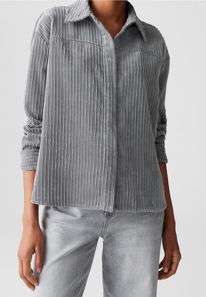 Kobieta w jasnoszarej prążkowanej koszuli sztruksowej z długimi rękawami podwiniętymi oraz w wyblakłych szarych dżinsach na jednolitym tle.