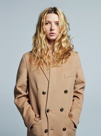 Manteau croisé en laine couleur chameau, doté de grands boutons foncés, d'une poche à rabat et d'une finition texturée, porté avec les mains dans les poches.