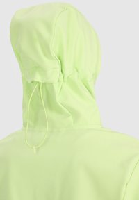 Personne portant une veste à capuche vert clair avec un cordon, vue de dos sur un fond uni clair.