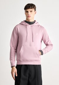 Soe roosa kapuutsiga sviiter, mis on valmistatud pehmest kangast, ees oleva tasku, paelaga kapuutsi ja väikese logoga plaastriga rinnal.