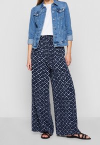 Mujer vestida con una chaqueta de mezclilla azul, una camiseta blanca, pantalones de pierna ancha de rayas azul marino y sandalias negras, de pie frente a un fondo simple.