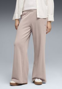 Personne portant un pantalon beige à jambes larges, des baskets blanches et une chemise blanche sur un fond dégradé gris.