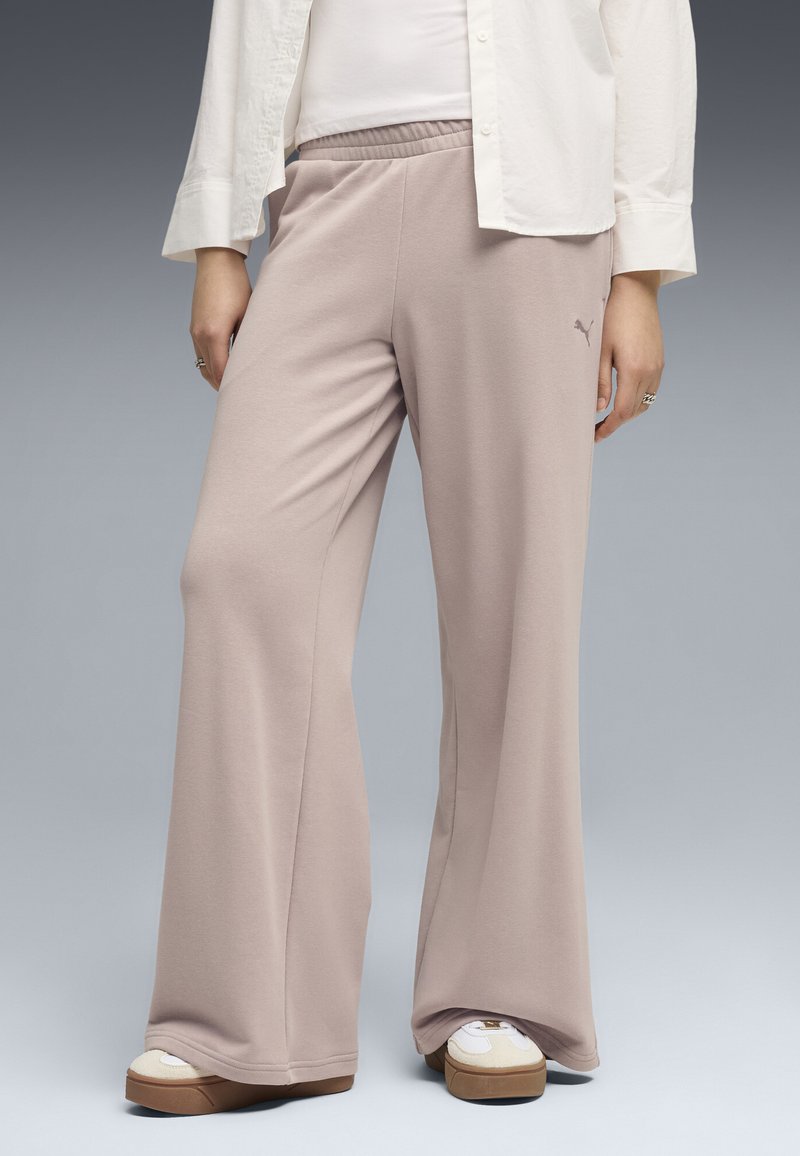 Personne portant un pantalon beige à jambes larges, des baskets blanches et une chemise blanche sur un fond dégradé gris.