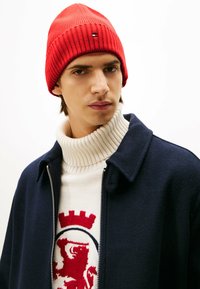 Rote, gerippte Strickmütze, kombiniert mit einem cremefarbenen Pullover mit hohem Kragen, der ein rotes Löwendesign zeigt, unter einem marineblauen Reißverschlussmantel.
