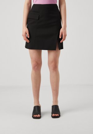 Femme portant une mini-jupe noire avec une fente latérale et une poche à rabat, des sandales noires ouvertes à l'avant, et un haut rose clair.