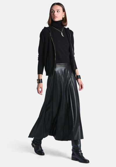 Femme portant un pull à col roulé noir, une veste à fermeture éclair, une longue jupe en cuir plissée, des bottes noires, des bijoux en or, posant avec un pied en avant.