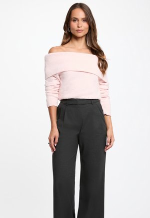 Kvinde iført en lyserød off-shoulder sweater og sorte, højtaljede bukser med vide ben, stående mod en hvid baggrund.
