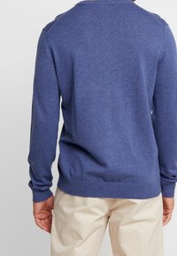 Niebieski sweter dziany z długimi rękawami, ściągaczami na mankietach i dole, gładką fakturą, okrągłym dekoltem, noszony na jasnych spodniach.