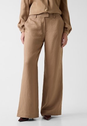 Beżowe spodnie o szerokich nogawkach zestawione z pasującą beżową bluzką i ciemnymi czółenkami na neutralnym tle.