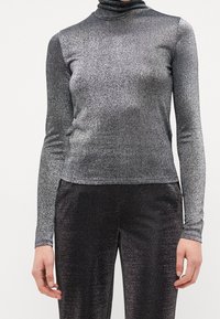 Metallisk silverfärgad polo med långa ärmar, gjord av ett stretchigt, glittrande tyg; har en figurnära silhuett och en slät textur.