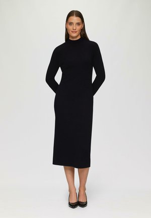 Robe en tricot noire arrivant aux genoux avec un col haut, des manches longues et une silhouette ajustée. Portée avec des escarpins noirs à bouts pointus. Texture lisse.