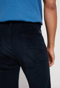 Merihenkiset corduroy-housut, joissa on teksturoitu pinta, takatasku ja pieni logo-patch vyötärön lähellä. Housut osittain näkyvissä.