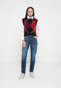 Adīta argaila stilā veste zilā, sarkanā un baltā krāsā virs baltas pogājamas krekla, kopā ar zilām džinsu biksēm un bēšām sporta kurpēm.