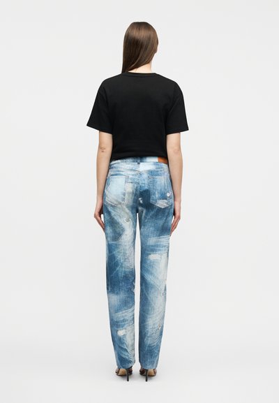 Γυναίκα που στέκεται με την πλάτη προς τα εμπρός, φοράει μαύρο πουκάμισο με κοντά μανίκια, ξεβαμμένα μπλε τζιν με φθορές και μαύρα σανδάλια με ψηλό τακούνι σε λευκό φόντο.