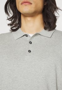 Vaaleanharmaa polo-paita, jossa on kauluskäänne, kolme mustaa nappia ja ribbikuvio. Paita on sopiva ja yksinkertainen muotoilultaan.