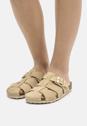 Scarpe slip-on in suede beige con design intrecciato, caratterizzate da una fibbia dorata e suola testurizzata. Punta e tallone aperti per una maggiore traspirabilità.