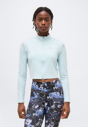 Νεαρή γυναίκα με πλεξούδες φοράει ανοιχτό μπλε κοντό μπουφάν με φερμουάρ και μαύρο κολάν ψηλόμεσο με μπλε και λευκό λουλουδάτο σχέδιο.