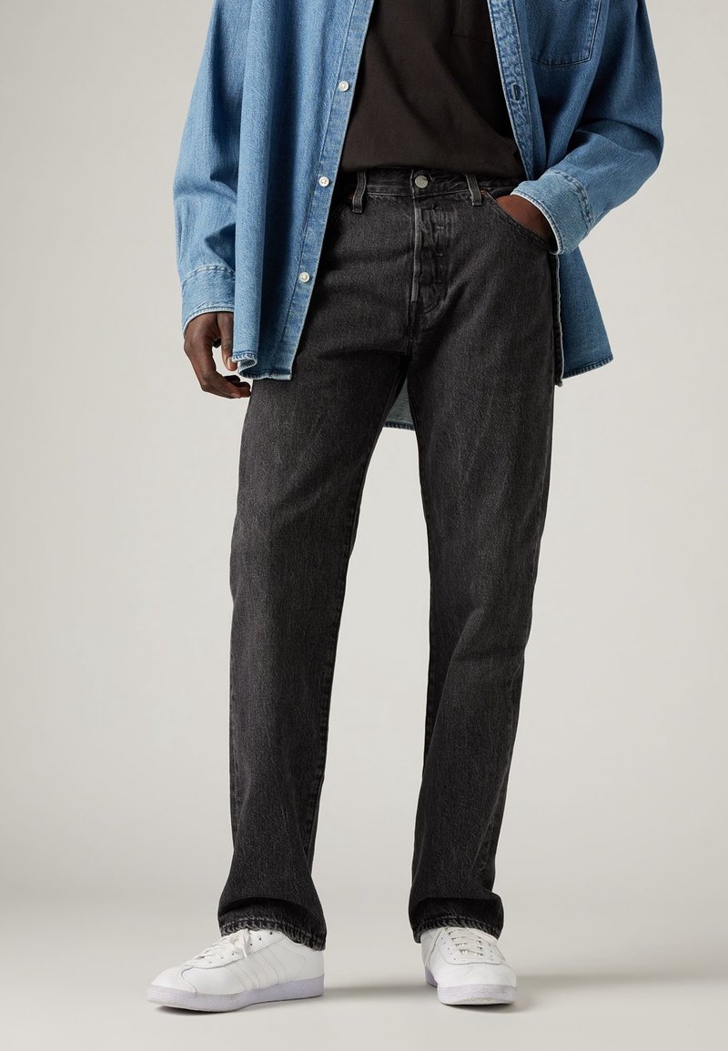 Personne portant un jean noir droit, des baskets blanches, une chemise noire et une veste en jean bleu déboutonnée avec la main dans la poche.