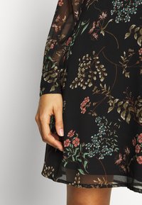 Schwarzes Kleid mit floralem Muster in gedeckten Farben, mit durchsichtigen langen Ärmeln und einem fließenden Saum, der florale Designs und Blätterakzente hervorhebt.