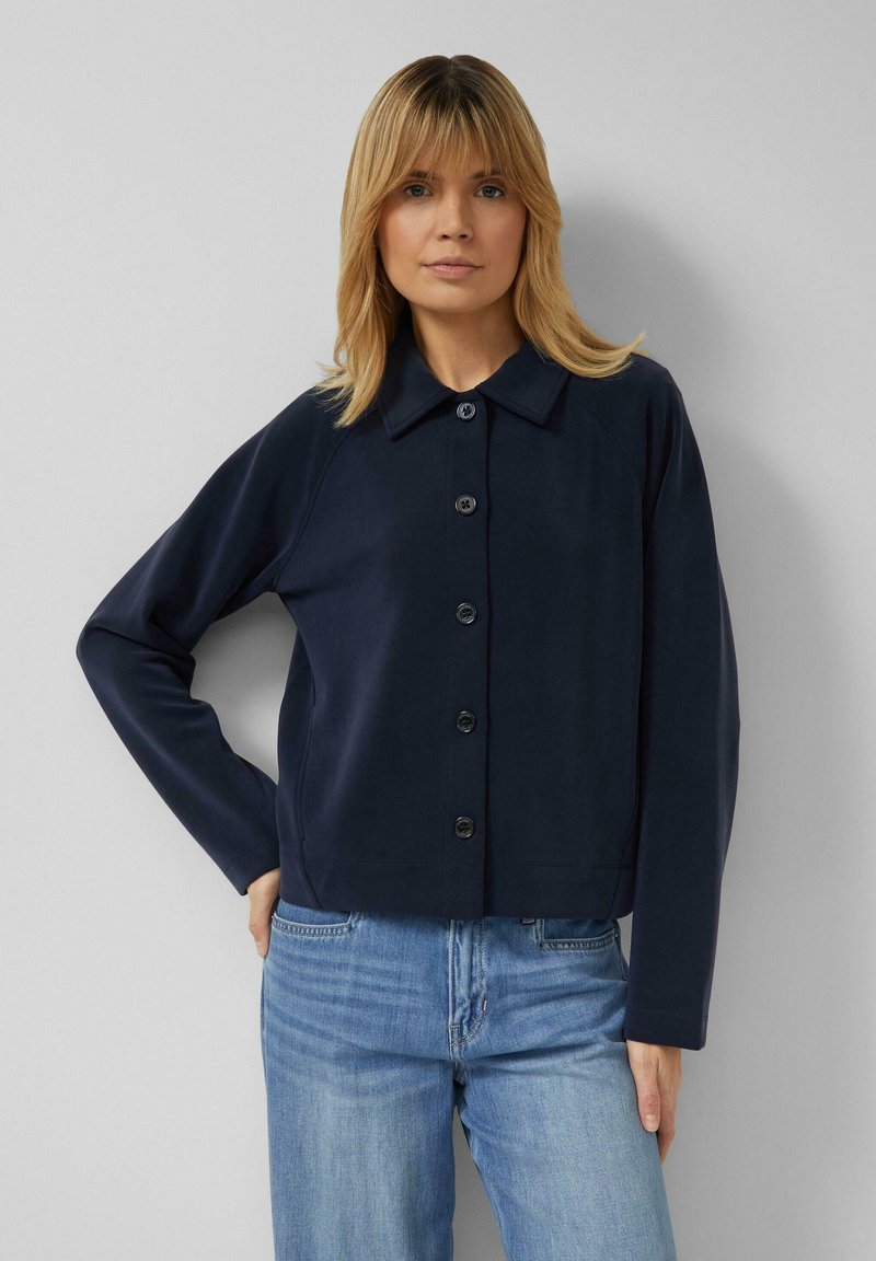 Frau mit schulterlangem blondem Haar, die eine marineblaue Jacke mit Knöpfen und hellblaue Jeans trägt, vor einem schlichten grauen Hintergrund stehend.