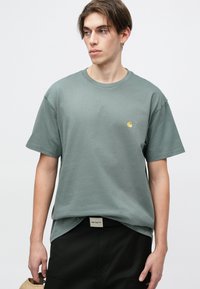 Olivgrünes Baumwoll-T-Shirt mit kurzen Ärmeln, Rundhalsausschnitt und einem kleinen goldenen Logo auf der linken Brust. Kombiniert mit dunklen Hosen und einem Gürtel.