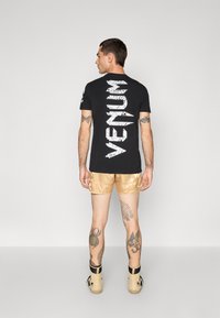 Schwarzes T-Shirt mit kurzen Ärmeln, das auf dem Rücken ein weißes, rissiges Logo zeigt, kombiniert mit glänzenden goldenen Shorts und hellfarbigem Schuhwerk.